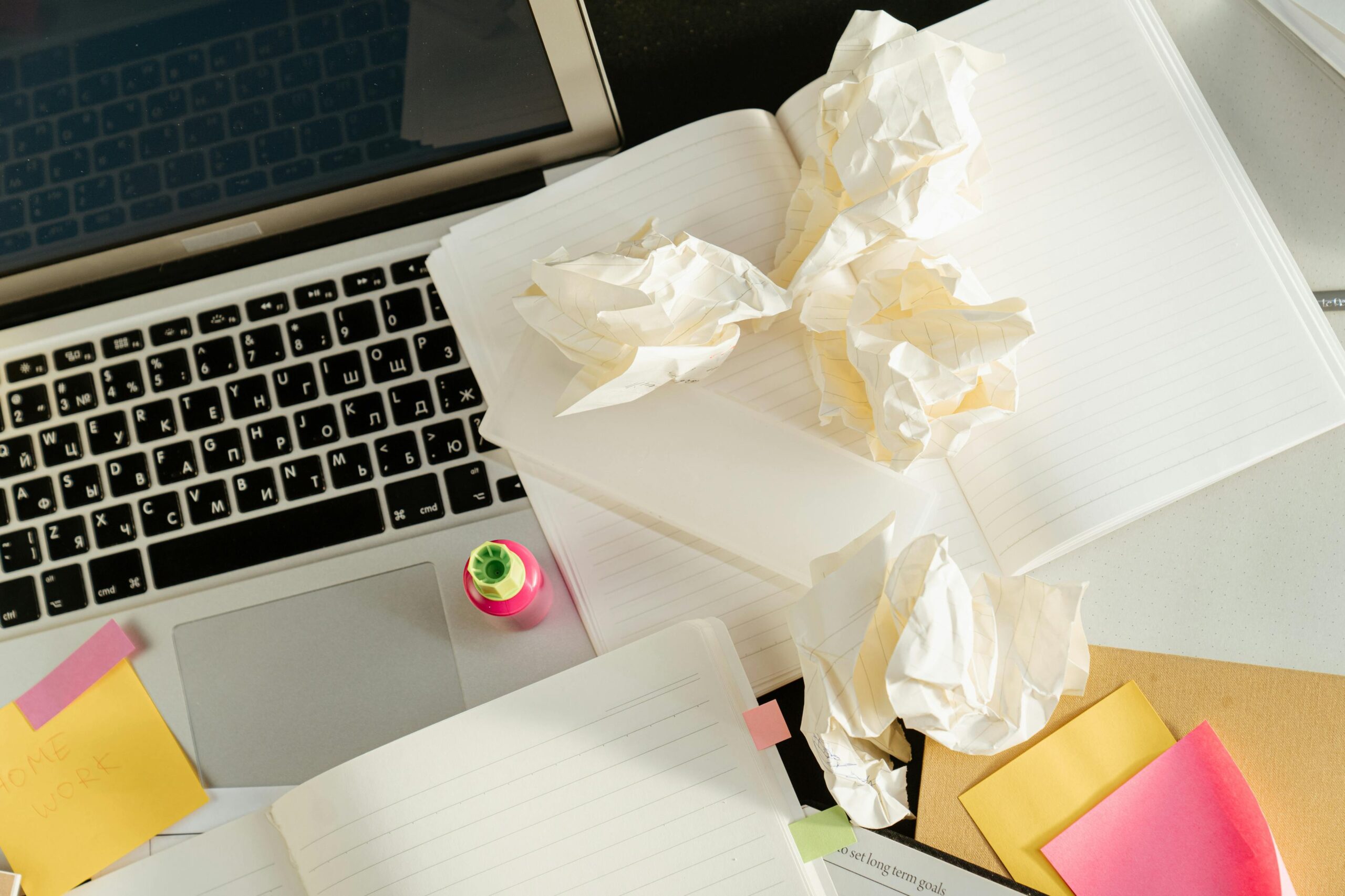A cluttered desk with laptop, notebooks, crumpled papers, and colorful sticky notes, showcasing creativity and brainstorming.